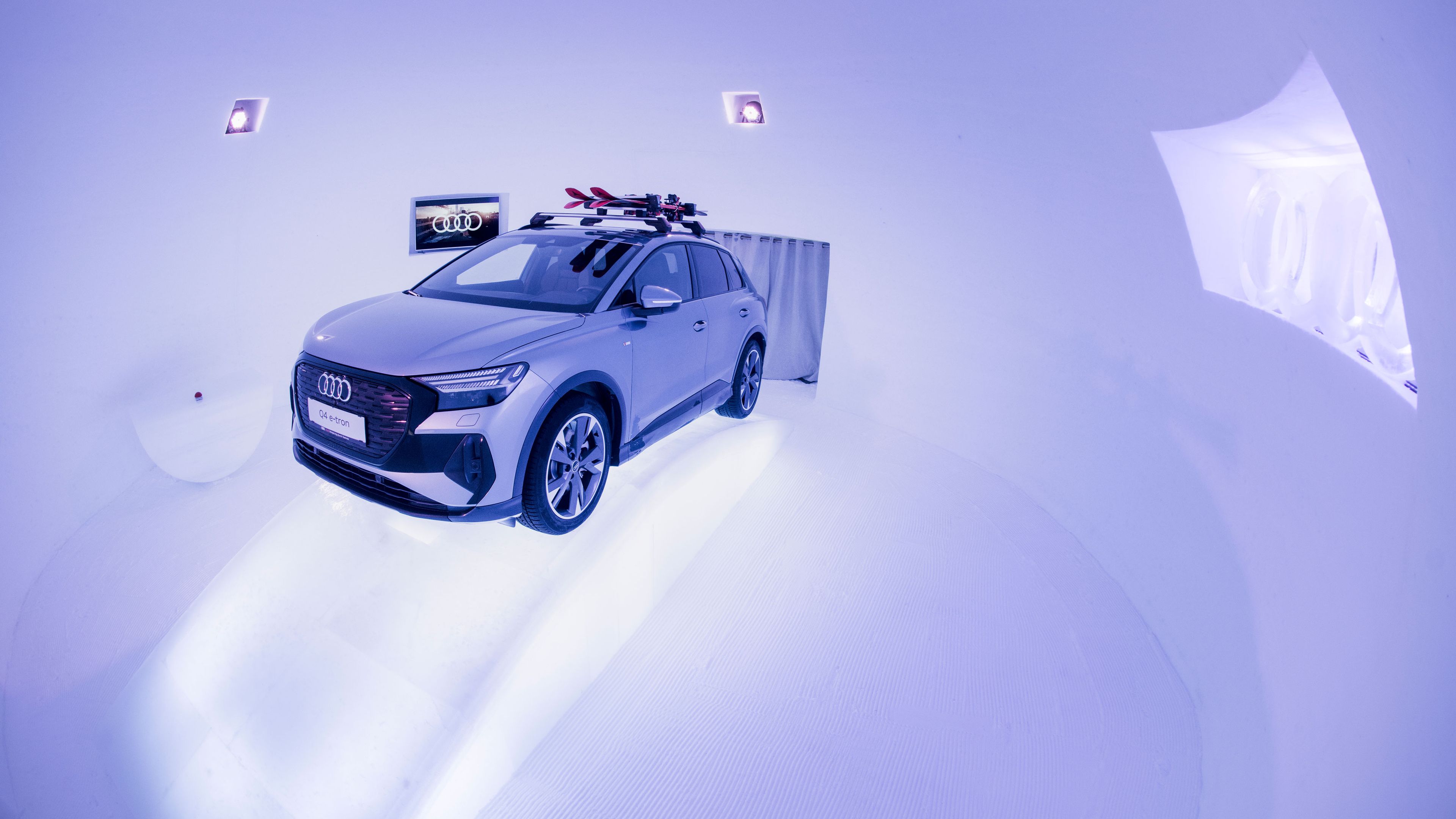 Ein silbergrauer Audi Q4 e-tron steht in einem futuristisch beleuchteten Raum aus Eis. Auf dem Dach befinden sich mehrere Ski. An der Wand hängt ein Bildschirm mit dem Audi-Logo.