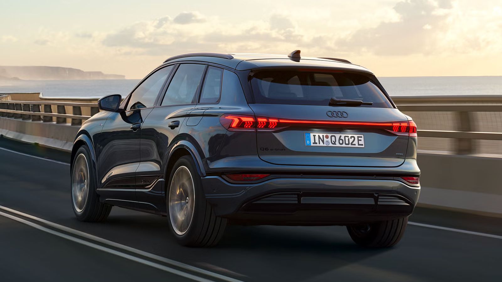 Schräge Heckaufnahme des Q6 SUV e-tron, welche über eine kurvige Straße am Meer fährt. Im Hintergrund sind Berge zu sehen.
