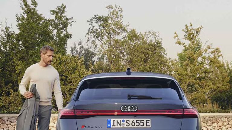 Heckprofil des S6 Avant e-tron, welcher auf Pflastersteinen vor einer Steinmauer und dem Garten eines modernen Hauses steht. Ein Mann mittleren Alters ist in Begriff, eine Tür zu öffnen.