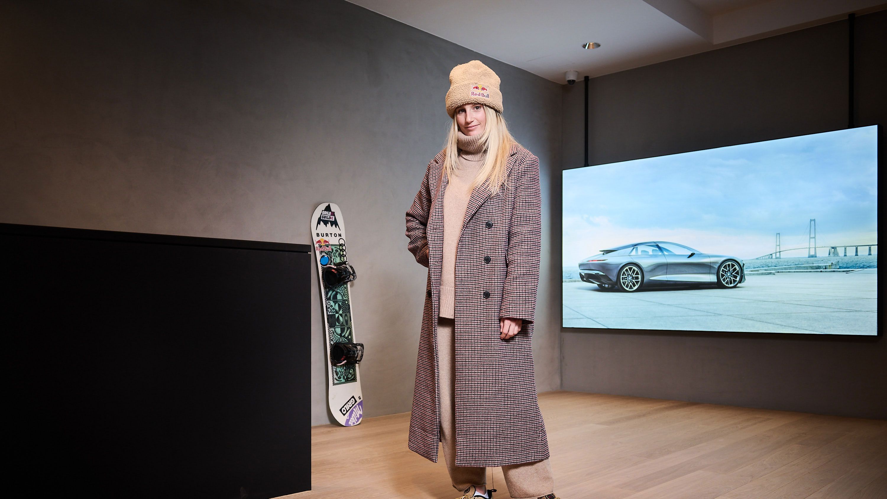 Anna Gasser in einem langen karierten Mantel und einer beige Mütze steht in einem modernen Raum. Hinter ihr ist ein großes Bild von einem Audi an der Wand und rechts daneben steht ein Snowboard auf dem Boden.