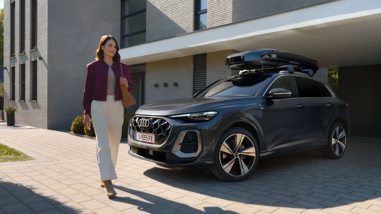 Frau passiert lächelnd einen in der Hauseinfahrt parkenden Audi Q5 mit Dachbox
