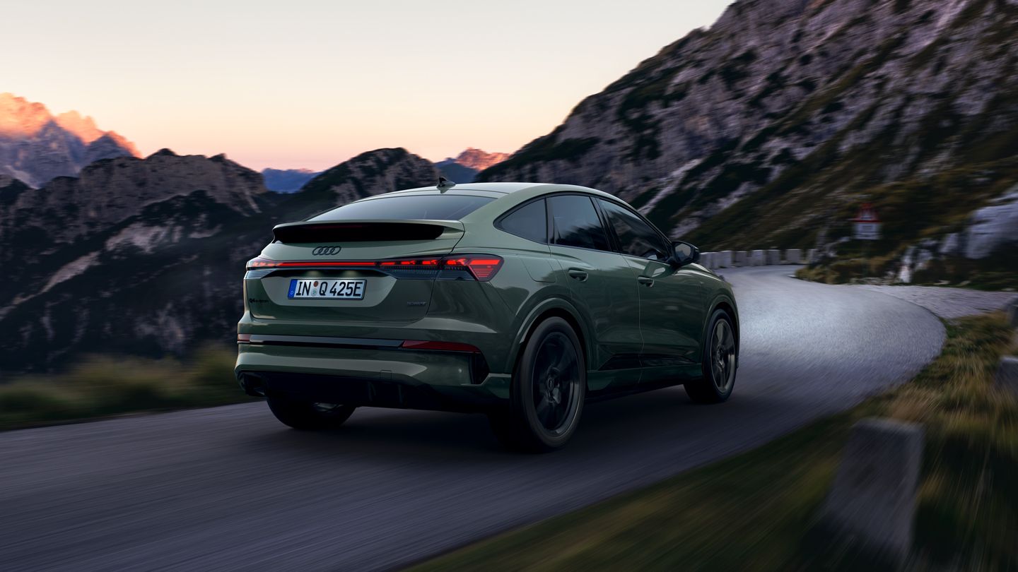 Audi Q4 Sportback e-tron fährt in der Abenddämmerung eine kurvige Bergstraße entlang. Im Hintergrund ragen steile Felsberge in den Himmel, deren Spitzen von der untergehenden Sonne leicht rosa beleuchtet werden.
