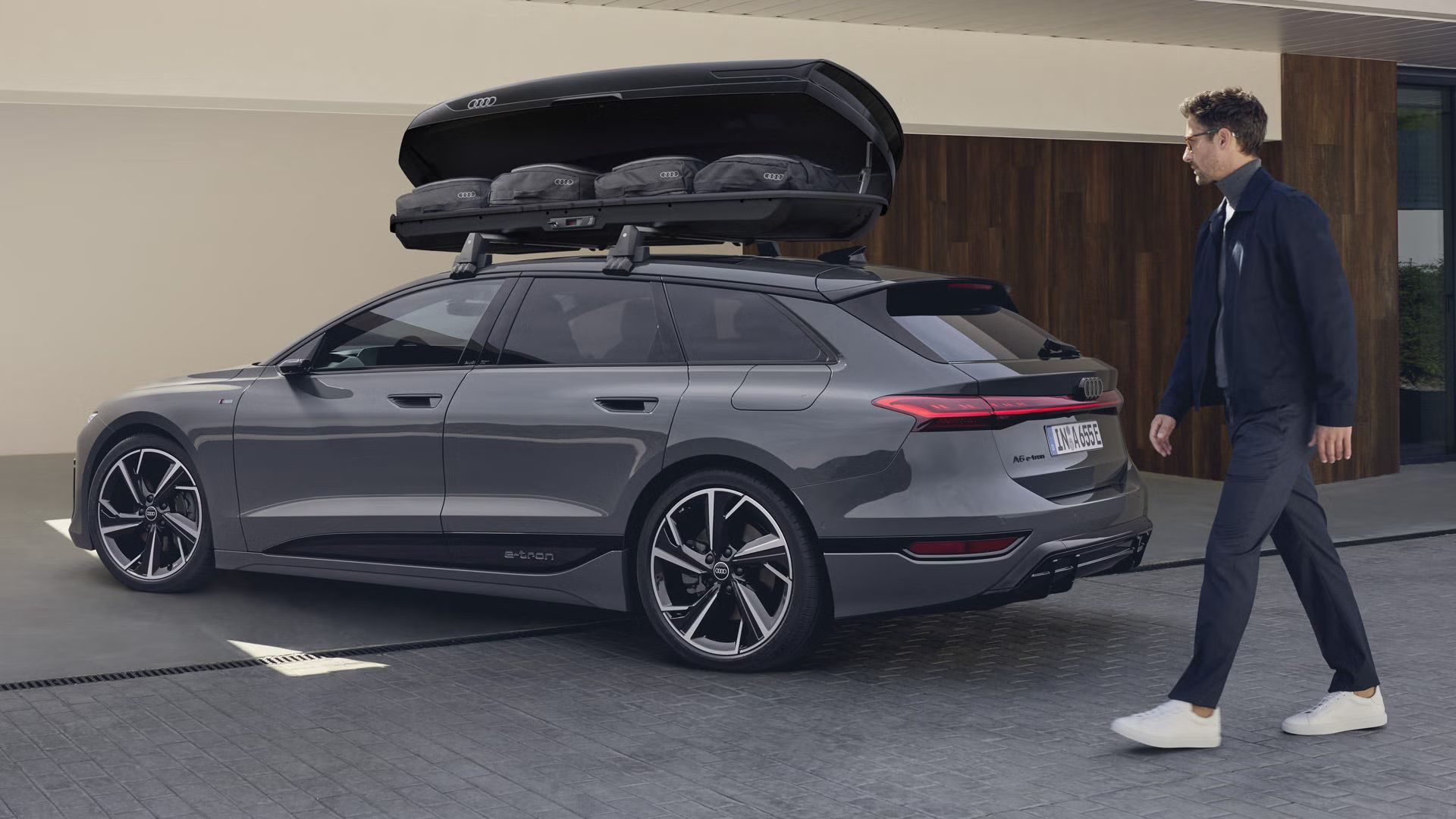 Der A6 Avant e-tron steht schräg vor einem modernen Haus, ein Mann bewegt sich auf das Fahrzeug zu. Auf dem Dach ist eine Dachbox montiert, deren Klappe geöffnet ist. Taschen sind in der Dachbox zu sehen. 