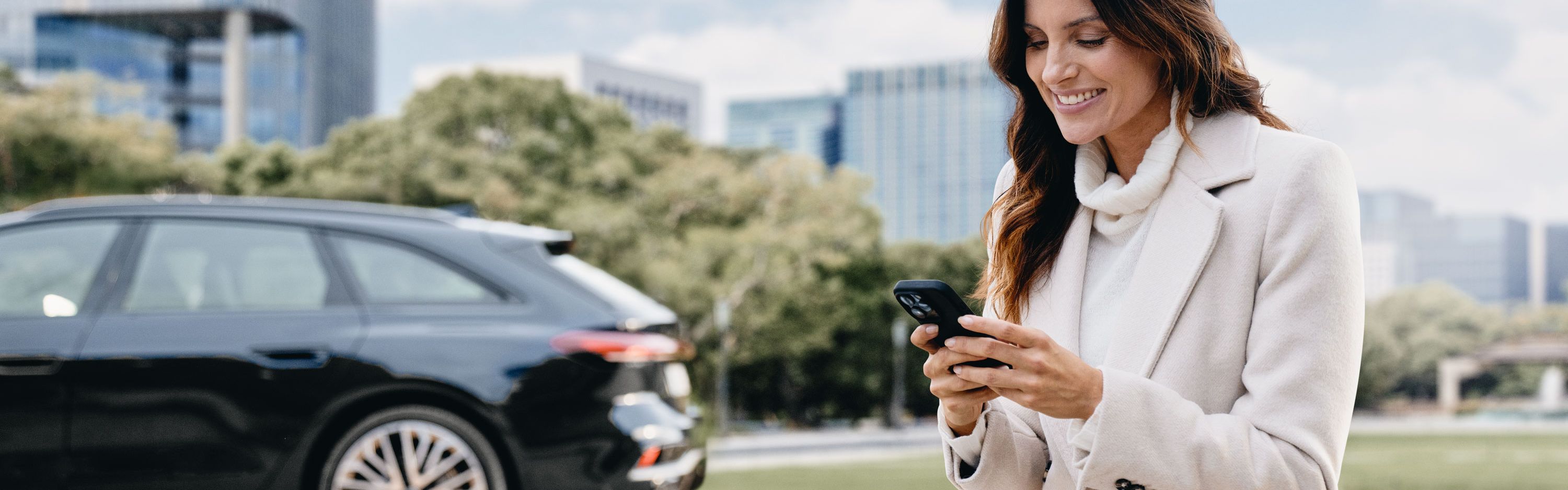Eine Frau sitzt im Freien auf einer Steinmauer und schaut lächelnd auf ihr Smartphone. Im Hintergrund ist ein schwarzer Audi vor modernen Bürogebäuden und Bäumen geparkt.