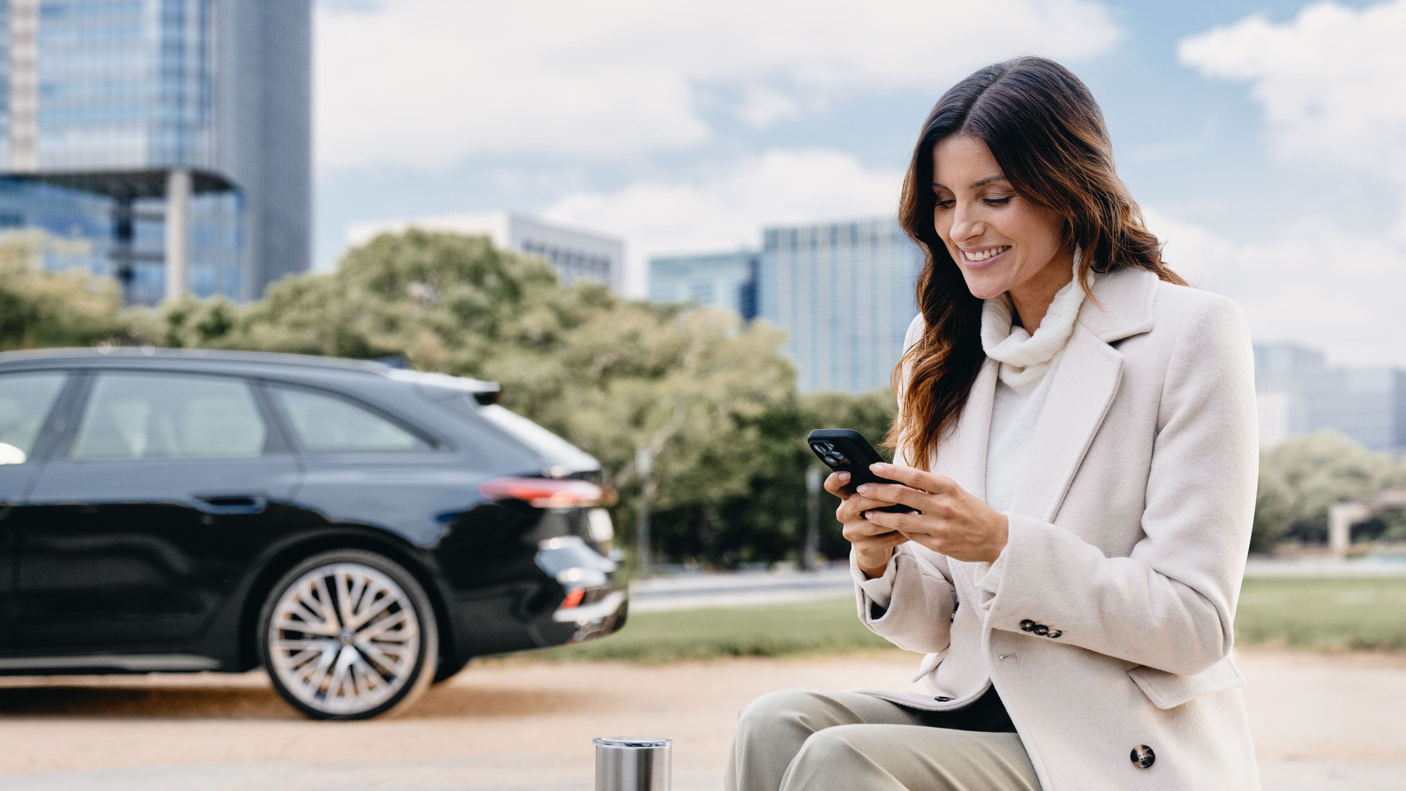 Eine Frau sitzt im Freien auf einer Steinmauer und schaut lächelnd auf ihr Smartphone. Im Hintergrund ist ein schwarzer Audi vor modernen Bürogebäuden und Bäumen geparkt.