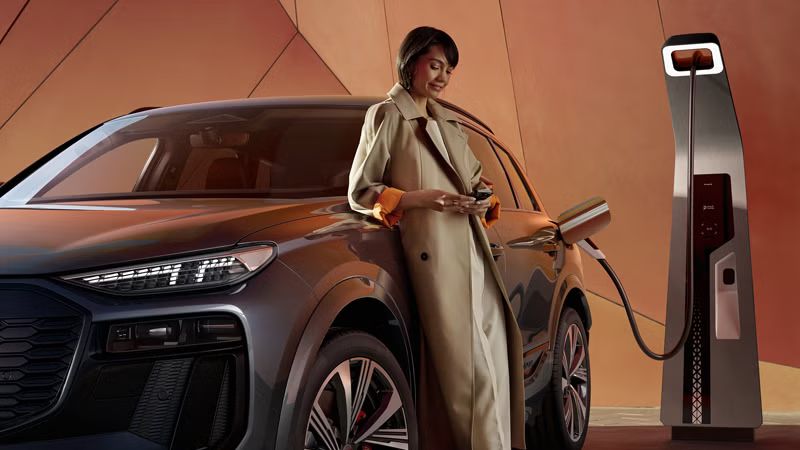Ein Audi SQ6 SUV e-tron wird an einer Ladesäule geladen. Auf dem Fahrzeug lehnt eine Frau.