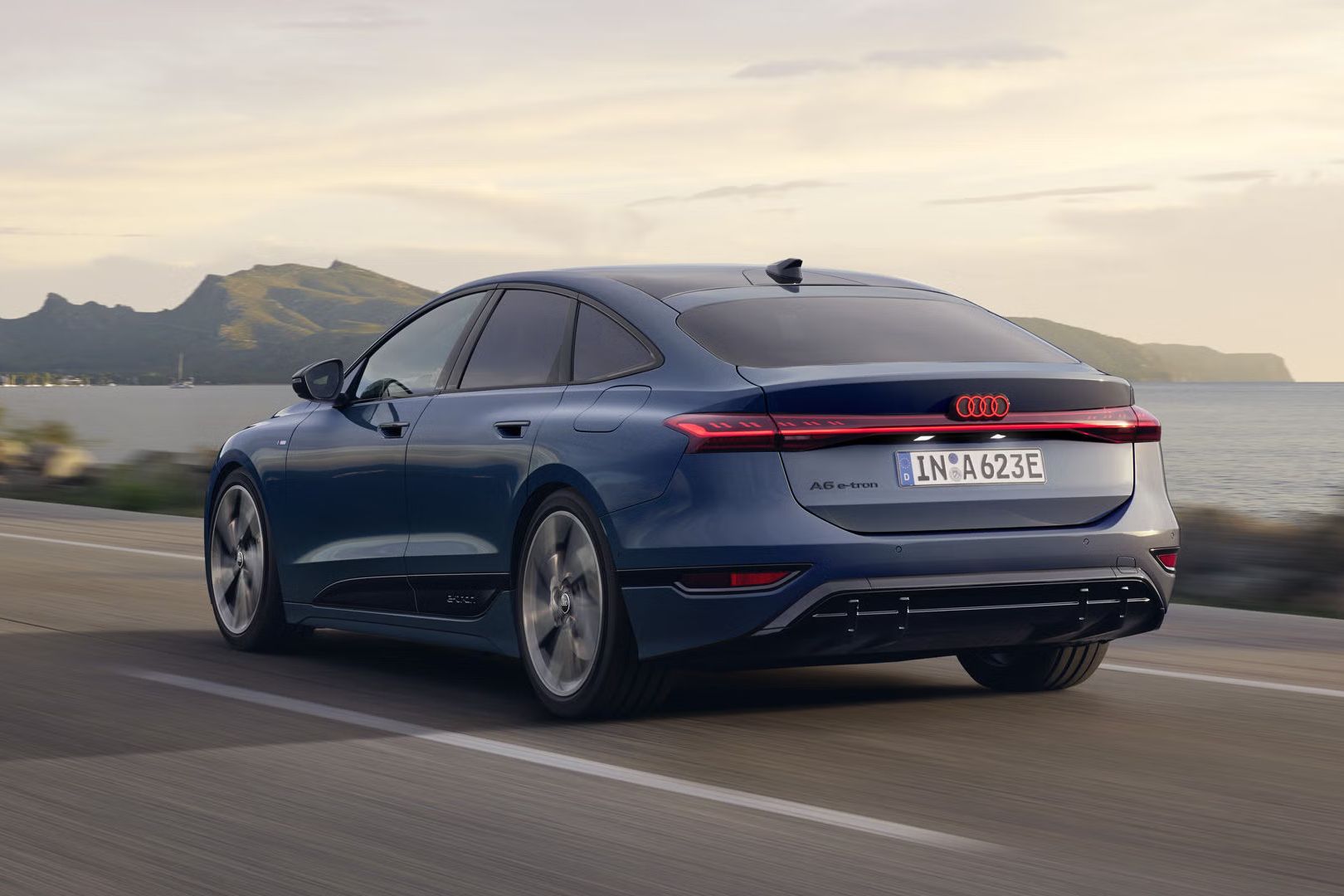 Der A6 Sportback e-tron aus schräger Heckansicht fährt eine Straße parallel zum Ufer entlang. Im Hintergrund sind Berge zu erkennen. 