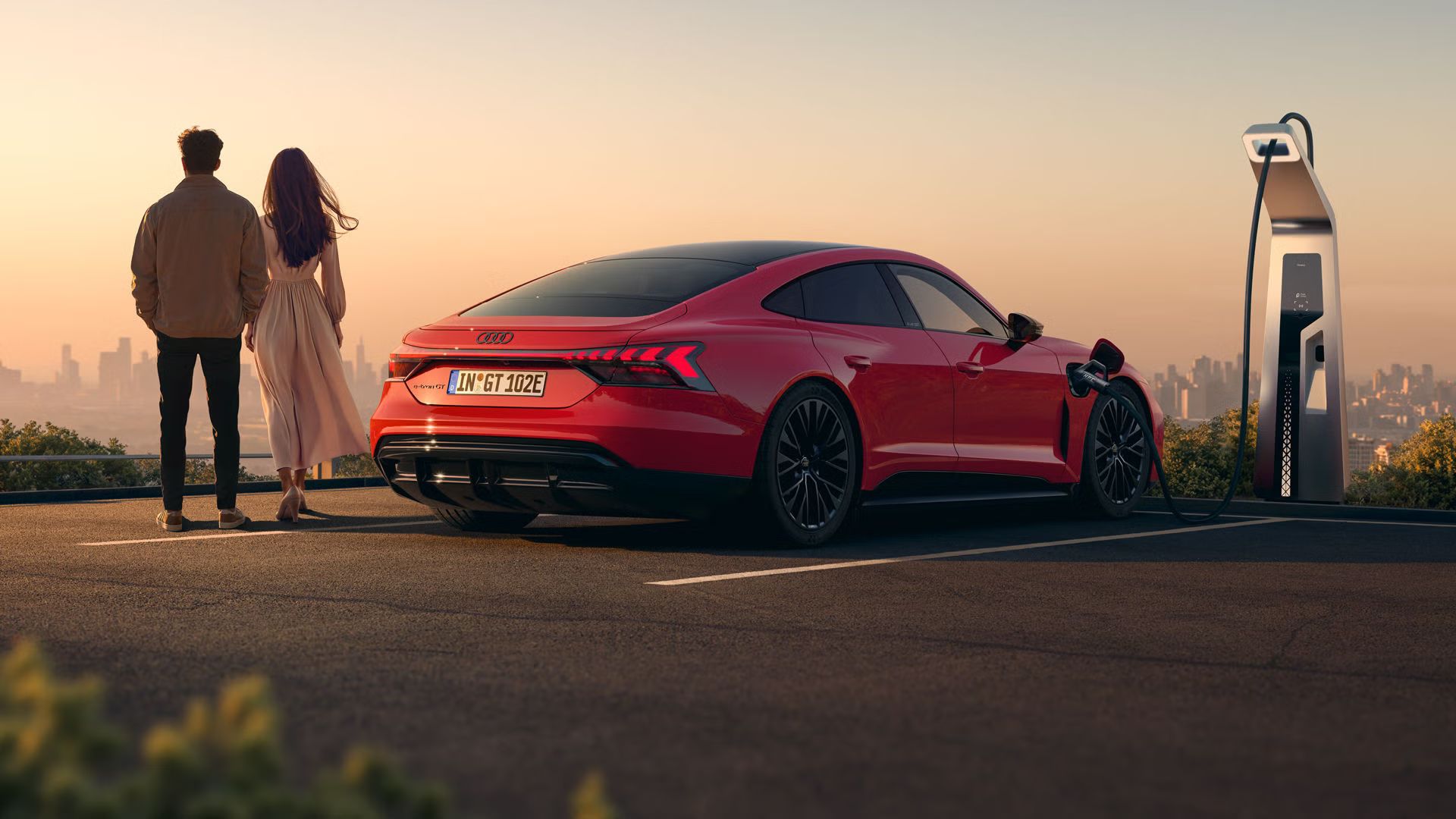 Ein roter Audi e-tron GT quattro steht auf einer Anhöhe über einer Stadt, die ins Licht eines Sonnenuntergangs gehüllt ist. Das Fahrzeug wird an einer öffentlicher Ladesäule geladen. Neben dem Fahrzeug stehen eine Frau und ein Mann.