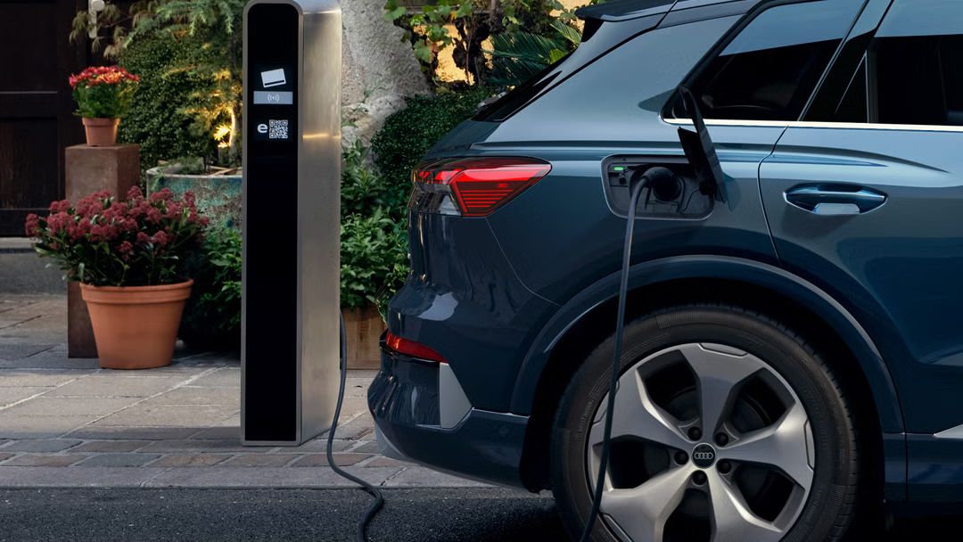 Nahaufnahme des rechten Hecks des Q4 SUV e-tron, parkend vor einem Gebäude, Pflanztöpfe sind vor dem Gebäude zu sehen. Das Fahrzeug ist an eine öffentliche Ladesäule angeschlossen. 