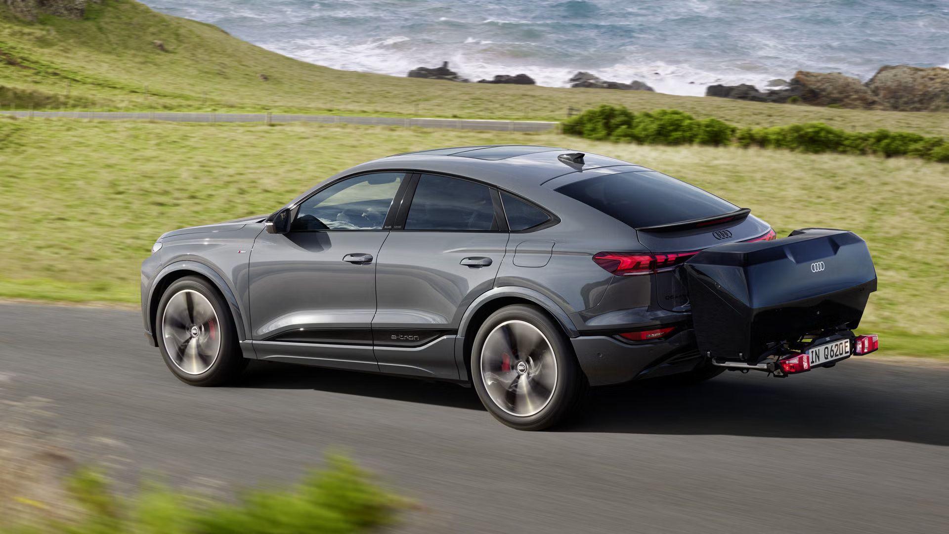 Der Q6 Sportback e-tron mit Heckbox fährt über eine Landstraße in der Natur.