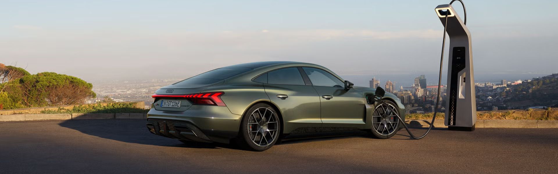 Grüner Audi RS e-tron GT performance lädt an einer Schnellladestation mit Stadtblick im Hintergrund.