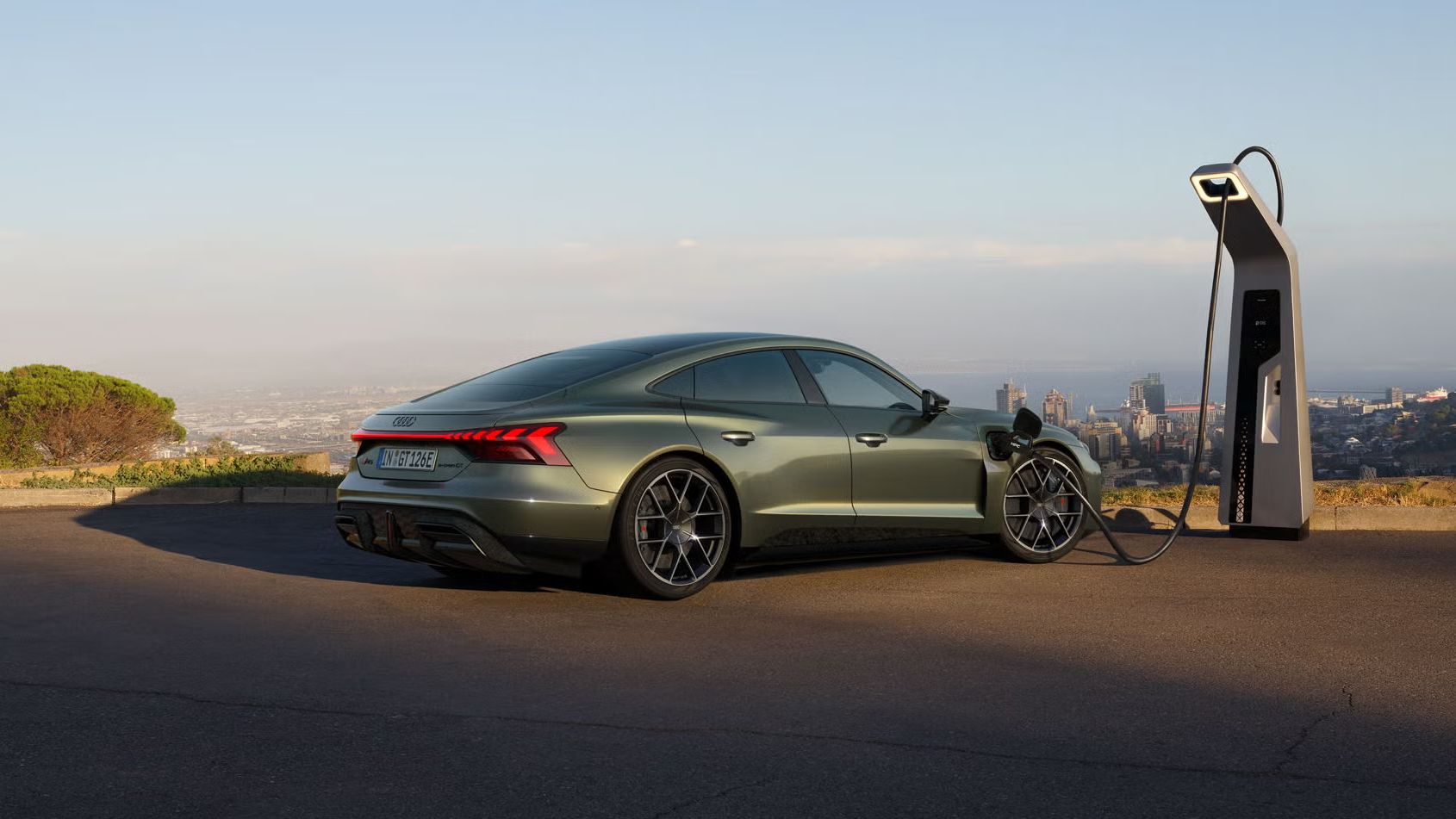 Grüner Audi RS e-tron GT performance lädt an einer Schnellladestation mit Stadtblick im Hintergrund.