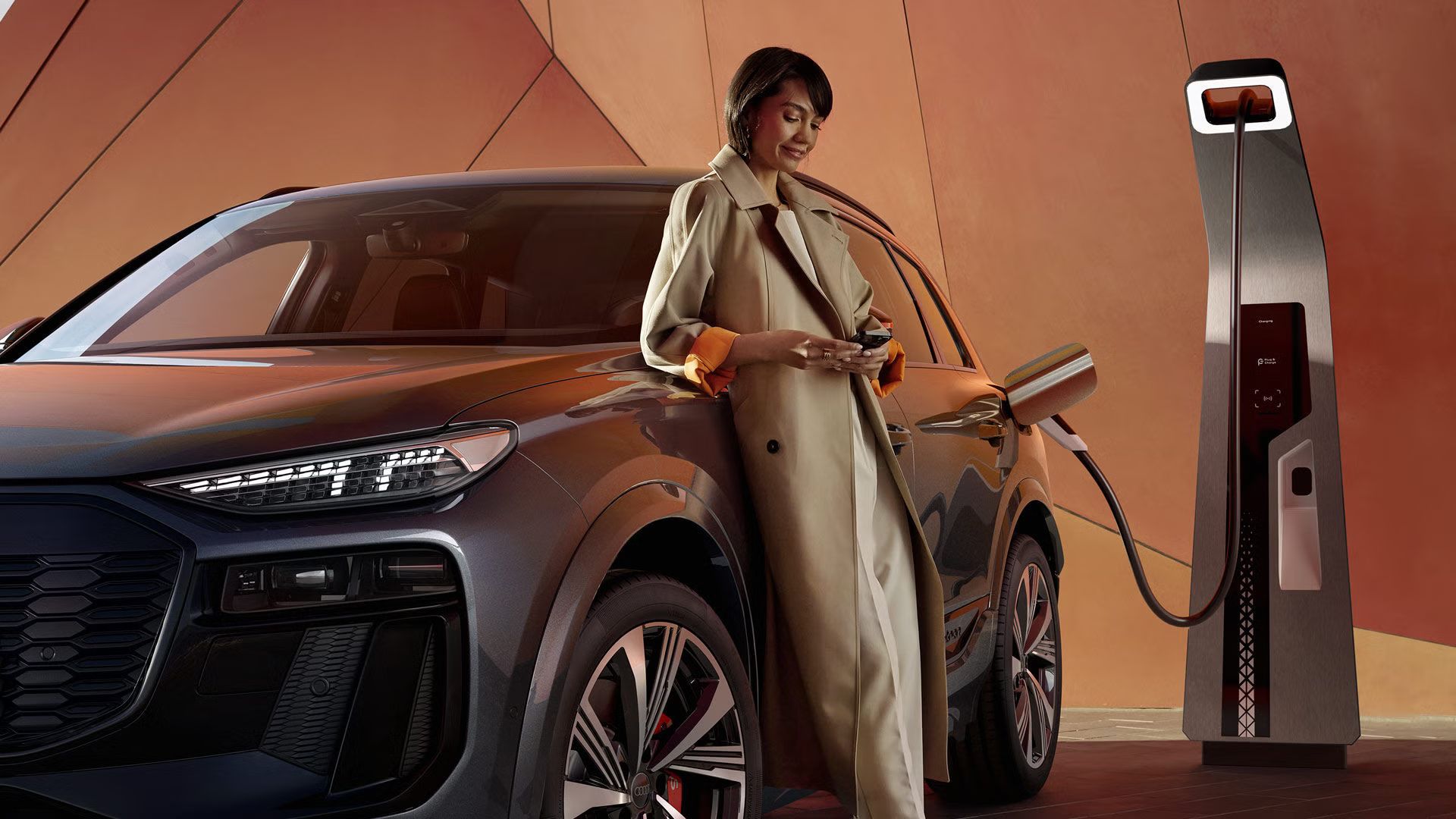 Ein Audi SQ6 SUV e-tron wird an einer Ladesäule geladen. Auf dem Fahrzeug lehnt eine Frau.