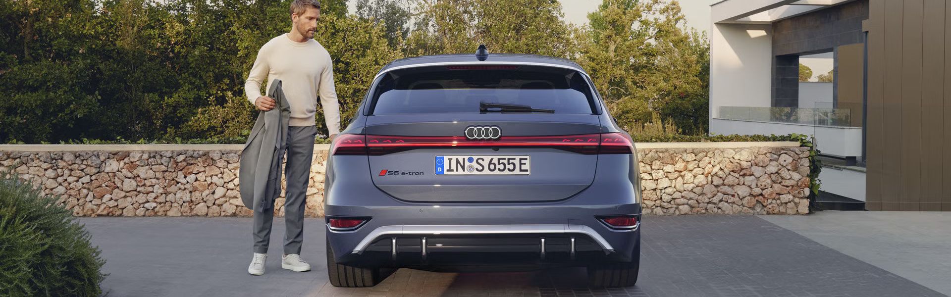 Heckprofil des S6 Avant e-tron, welcher auf Pflastersteinen vor einer Steinmauer und dem Garten eines modernen Hauses steht. Ein Mann mittleren Alters ist in Begriff, eine Tür zu öffnen.
