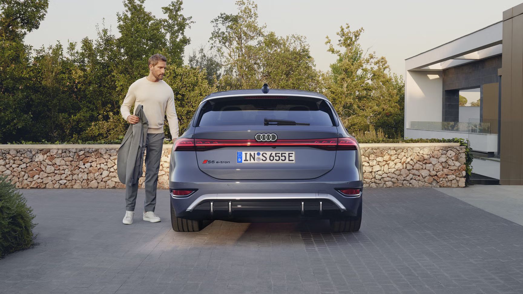 Heckprofil des S6 Avant e-tron, welcher auf Pflastersteinen vor einer Steinmauer und dem Garten eines modernen Hauses steht. Ein Mann mittleren Alters ist in Begriff, eine Tür zu öffnen.