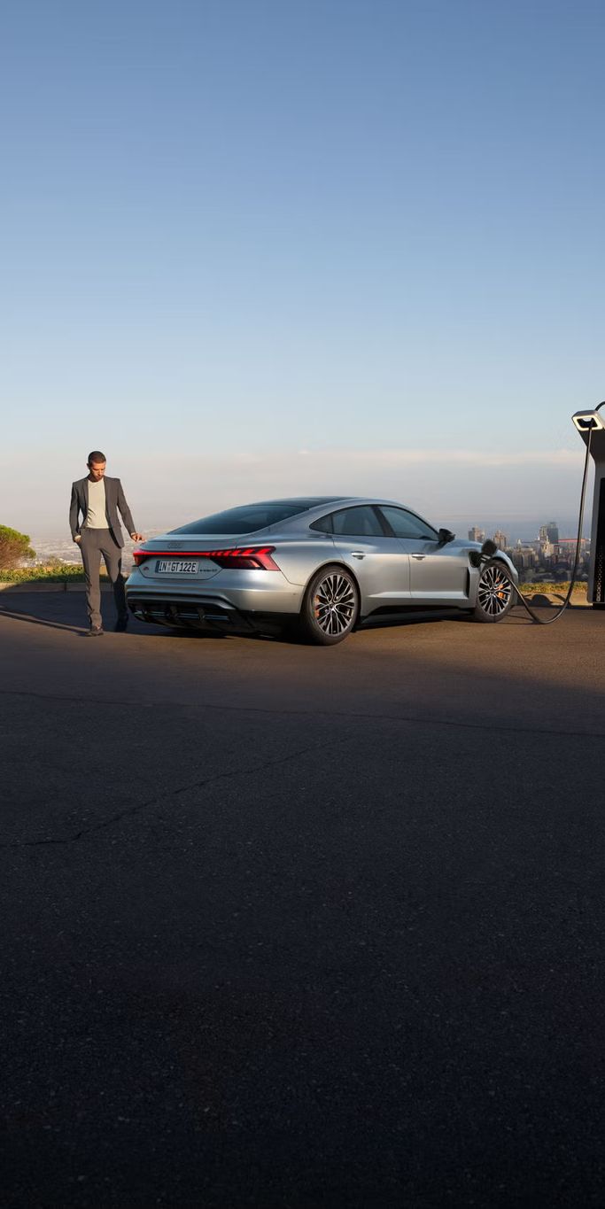 Ein silberner Audi S e-tron GT lädt an einer Ladestation mit Blick auf eine Stadt. Ein Mann im Anzug geht neben dem Auto entlang.