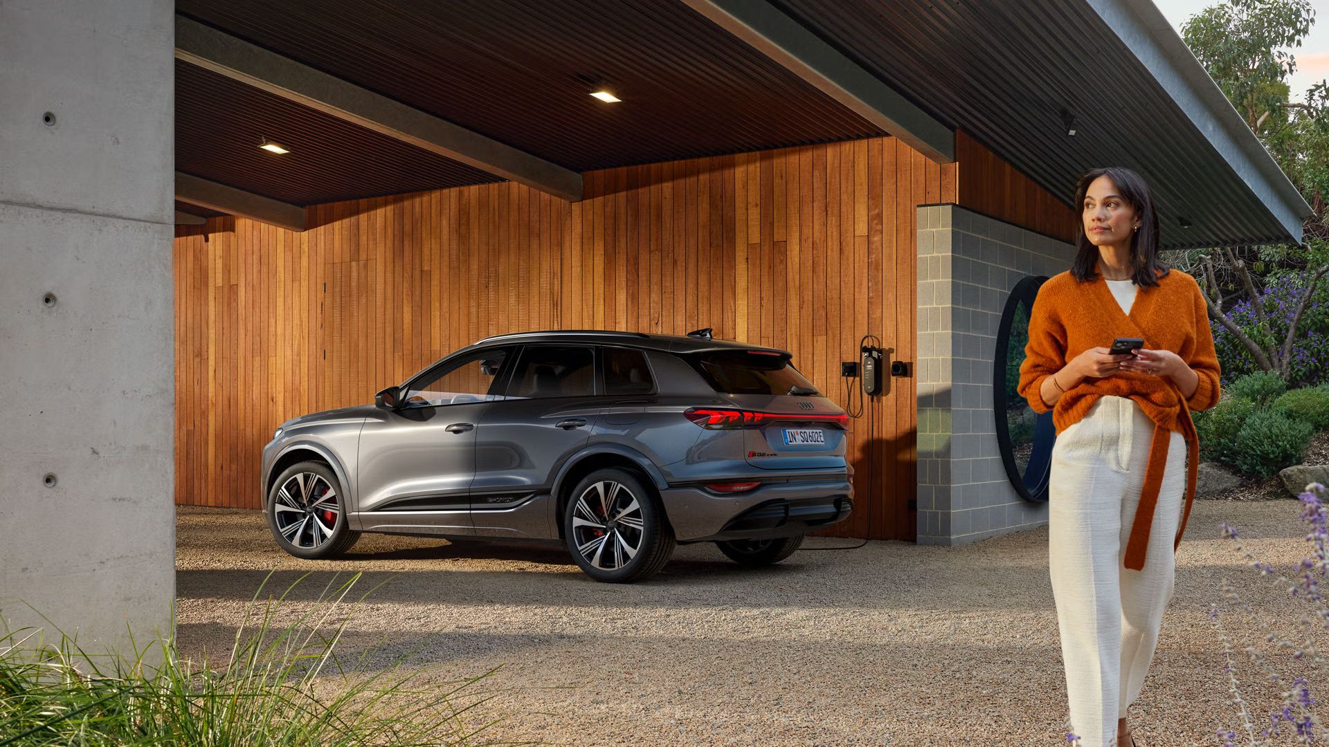 Seitenprofil des SQ6 SUV e-tron, welcher vor einer Holzhausfassade steht und an eine an der Hauswand montierte Ladestation angeschlossen ist.