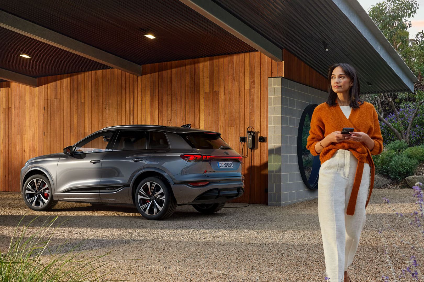 Seitenprofil des SQ6 SUV e-tron, welcher vor einer Holzhausfassade steht und an eine an der Hauswand montierte Ladestation angeschlossen ist.