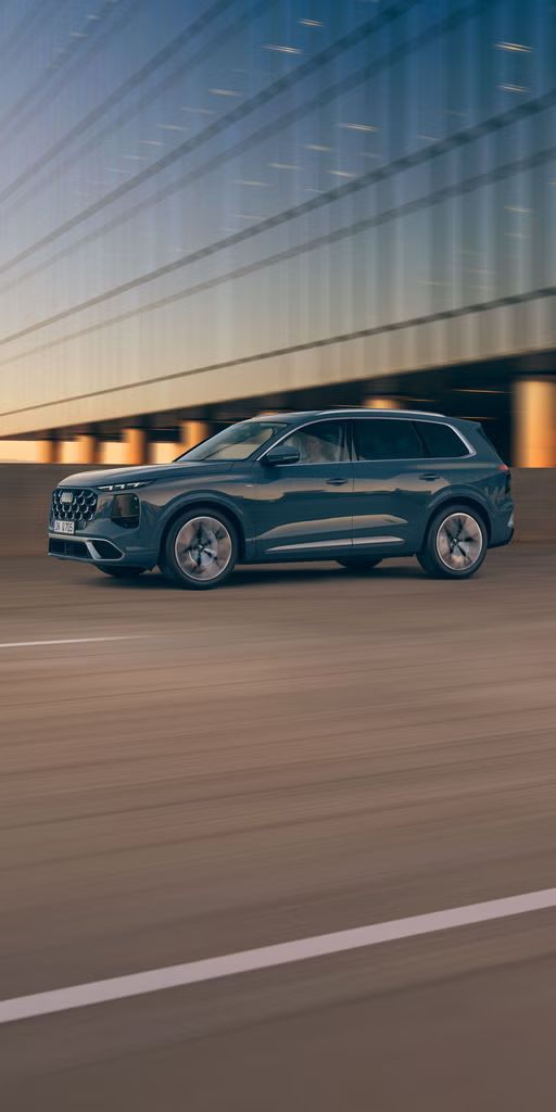 Ein blauer Audi Q7 SUV fährt auf einer Straße bei Sonnenuntergang, im Hintergrund ein modernes Gebäude auf Betonpfeilern.