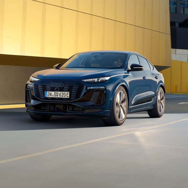 Schräges Frontprofil des SQ6 Sportback e-tron, welcher auf einer Straße durch die Stadt fährt. Im Hintergrund sieht man eine gelbe Gebäudefassade.