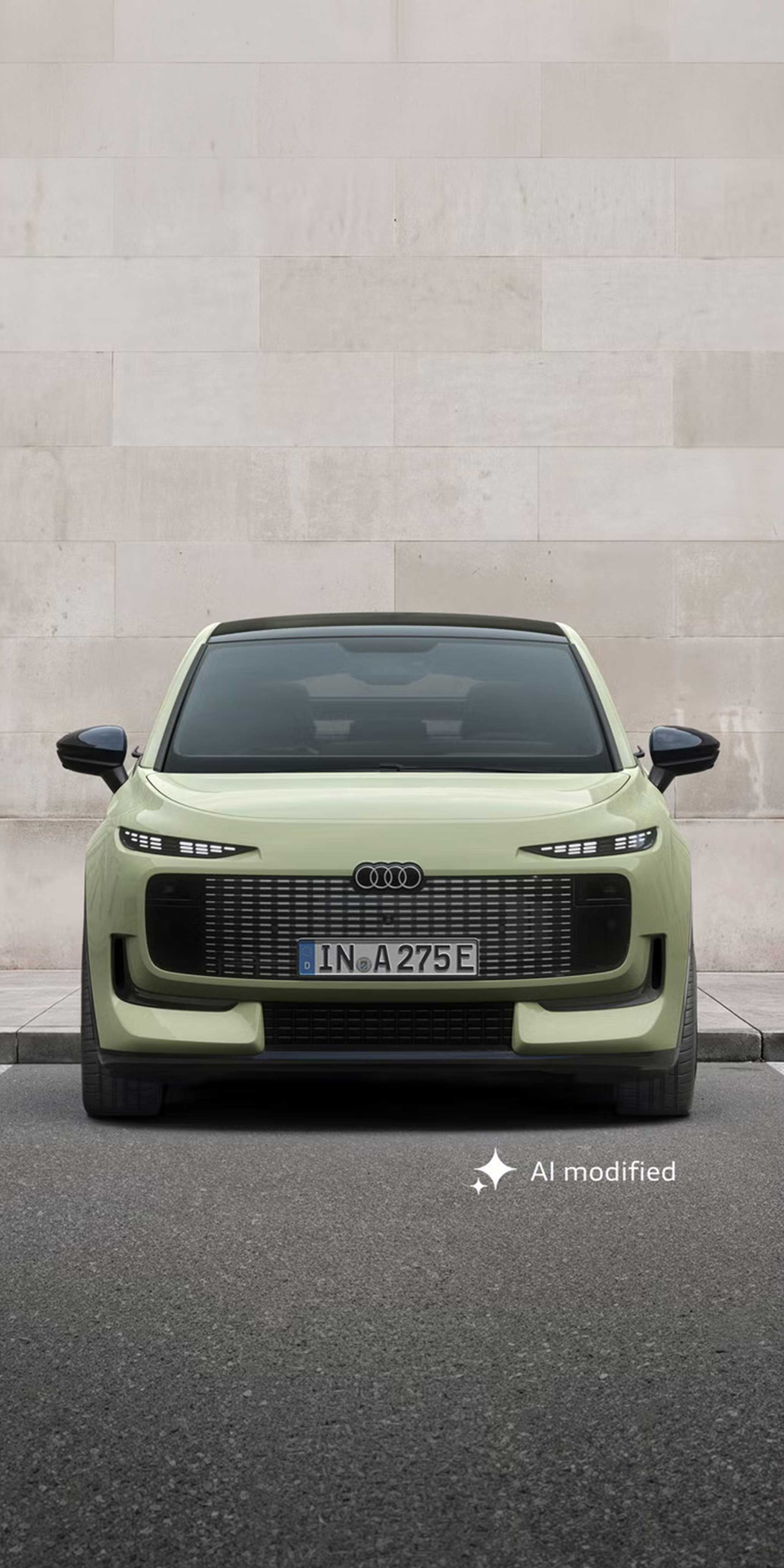 Der Audi A2 e-tron frontal von vorn auf einem Parkplatz, dahinter eine Gebäudewand in Steinoptik.