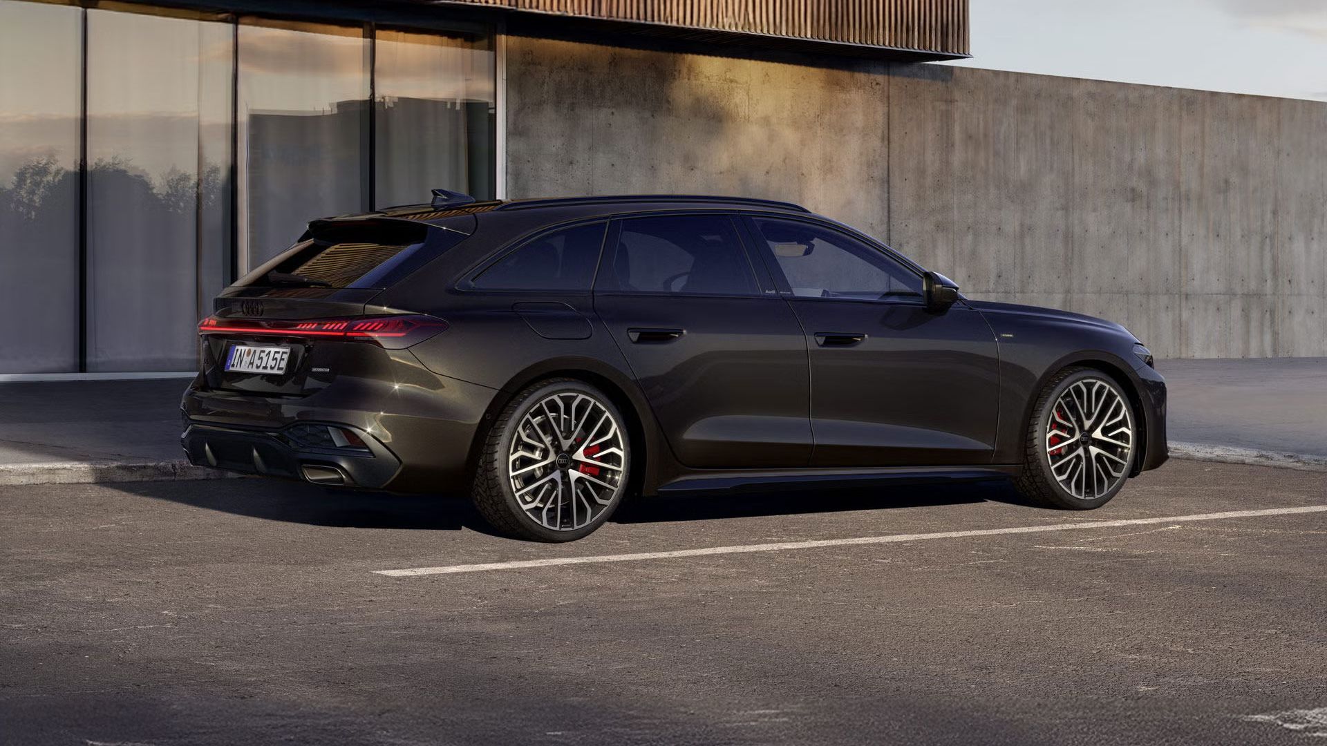 Der A5 Avant e-hybrid in der Lackierung Havannaschwarz Metallic steht im schrägen Seitenprofil auf einem Parkplatz vor einem Gebäude.