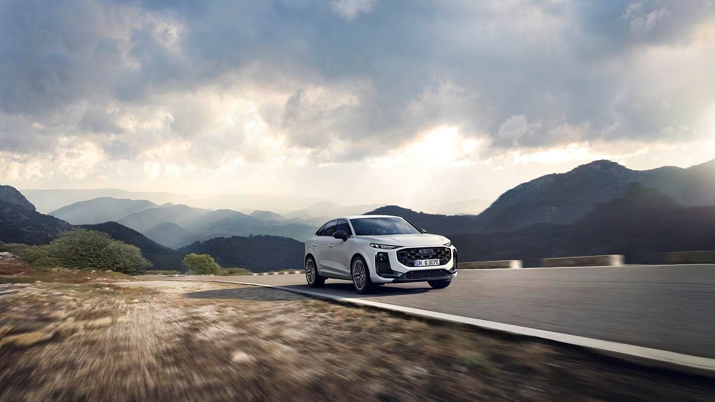 Ein Audi Q3 Sportback e-hybrid fährt eine bergige Landstraße entlang unter einem bewölkten Himmel. Die Landschaft ist weitläufig und zeigt Berge und Täler. 
