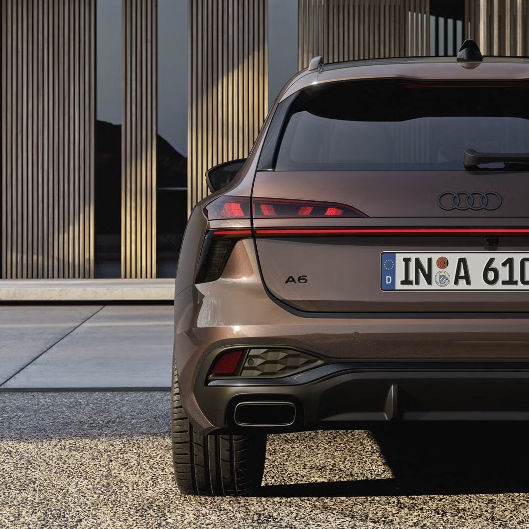 Heckaufnahme des A6 Avant, der auf einer Kiesfläche vor einer Fassade mit Holzvertäfelung und hohen Fenstern steht.