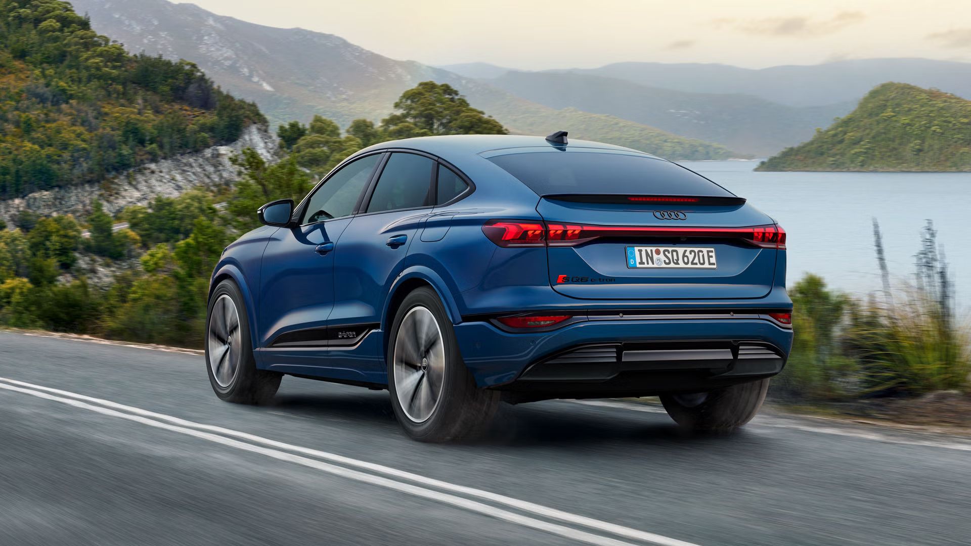 Schräge Heckaufnahme des SQ6 Sportback e-tron, welcher eine Straße an einem See in einer bergigen Landschaft entlangfährt.