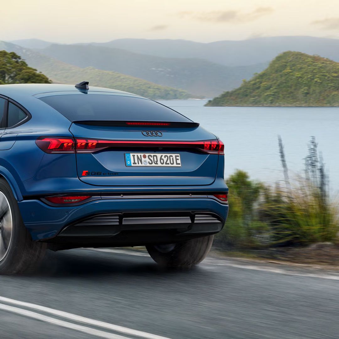 Schräge Heckaufnahme des SQ6 Sportback e-tron, welcher eine Straße an einem See in einer bergigen Landschaft entlangfährt.