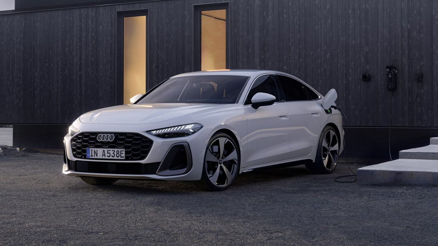 Die A5 Limousine e-hybrid im schrägen Frontprofil. Das Fahrzeug steht vor der dunklen Fassade eines Gebäudes und ist an eine Ladestation angeschlossen.