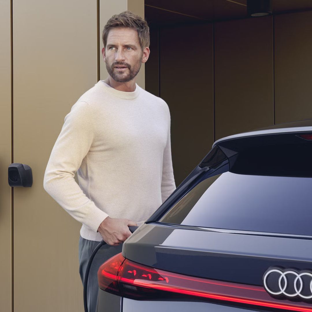 Der S6 Avant e-tron steht vor einem modernen Haus. An der Hausfassade ist eine Ladestation montiert, welche von einem Mann mittleren Alters an das Fahrzeug angeschlossen wird.