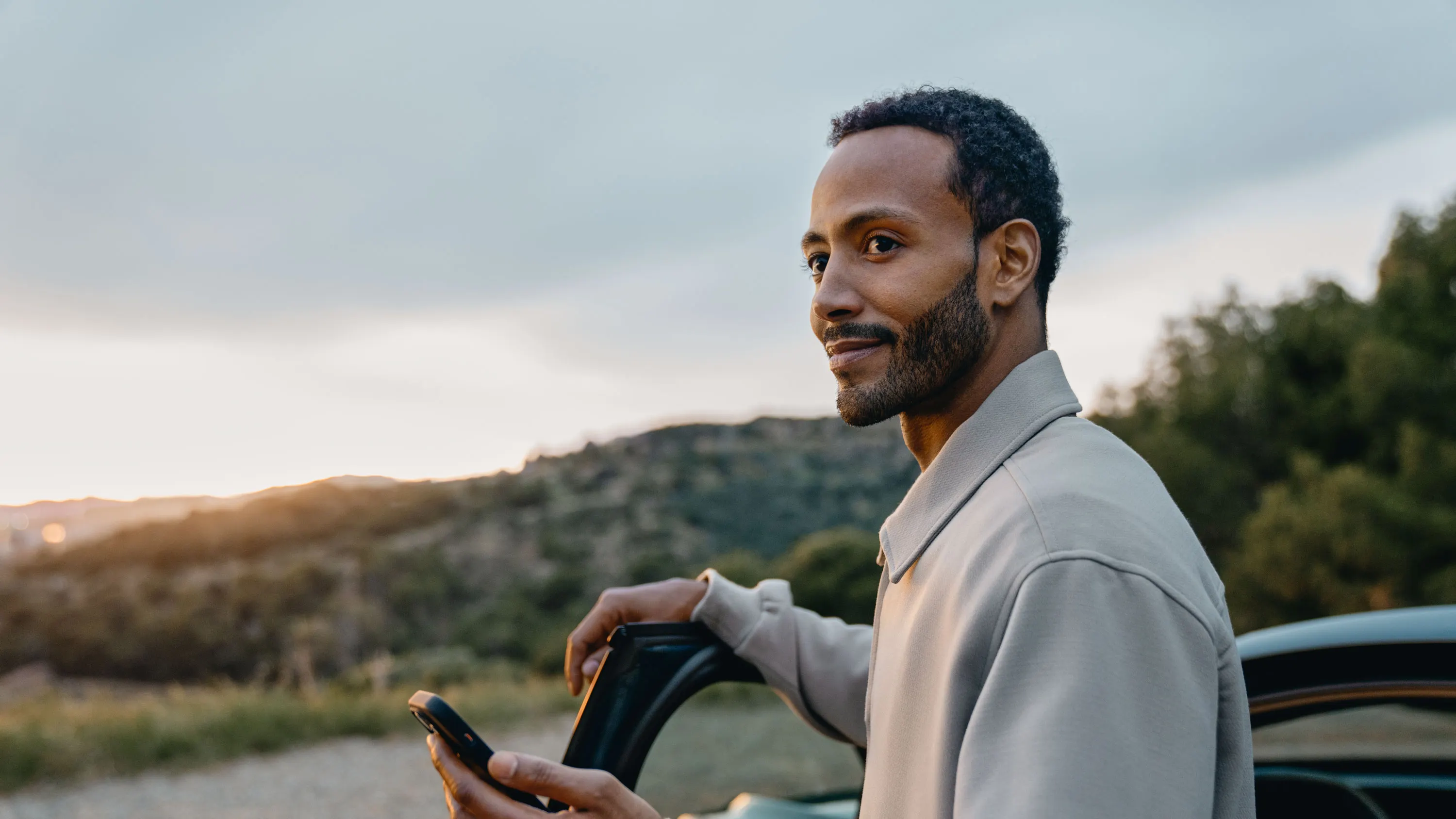 Ein junger Mann steht an der offenen Autotür und hält ein Smartphone in der Hand. Er blickt seitlich lächelnd in die Ferne, während im Hintergrund eine hügelige Landschaft bei Sonnenuntergang zu sehen ist.