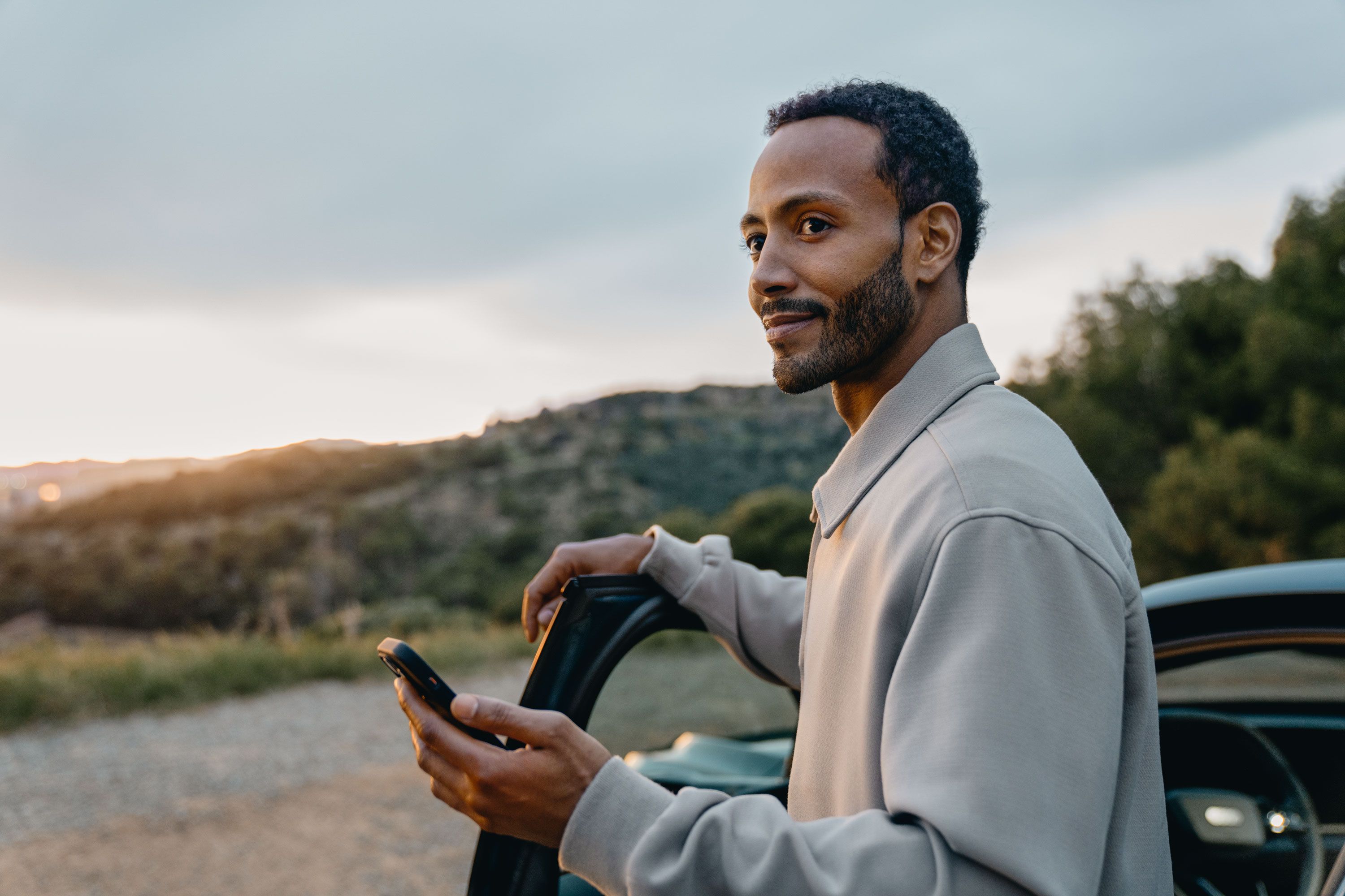 Ein junger Mann steht an der offenen Autotür und hält ein Smartphone in der Hand. Er blickt seitlich lächelnd in die Ferne, während im Hintergrund eine hügelige Landschaft bei Sonnenuntergang zu sehen ist.