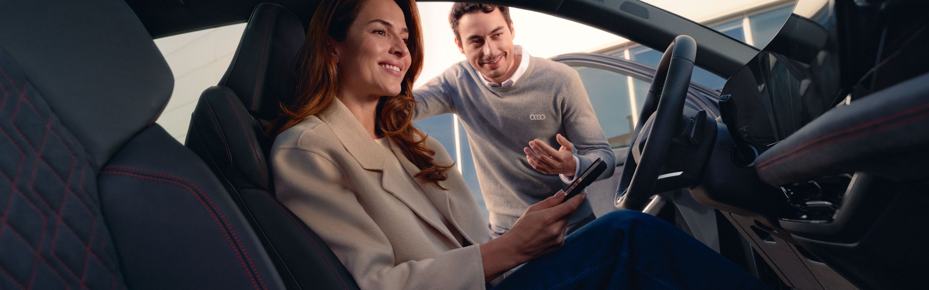 Eine Frau sitzt lächelnd auf dem Fahrersitz eines Audi und hält ein Smartphone in der Hand. Neben dem offenen Fahrzeug beugt sich ein Serviceberater herein und spricht freundlich mit ihr.