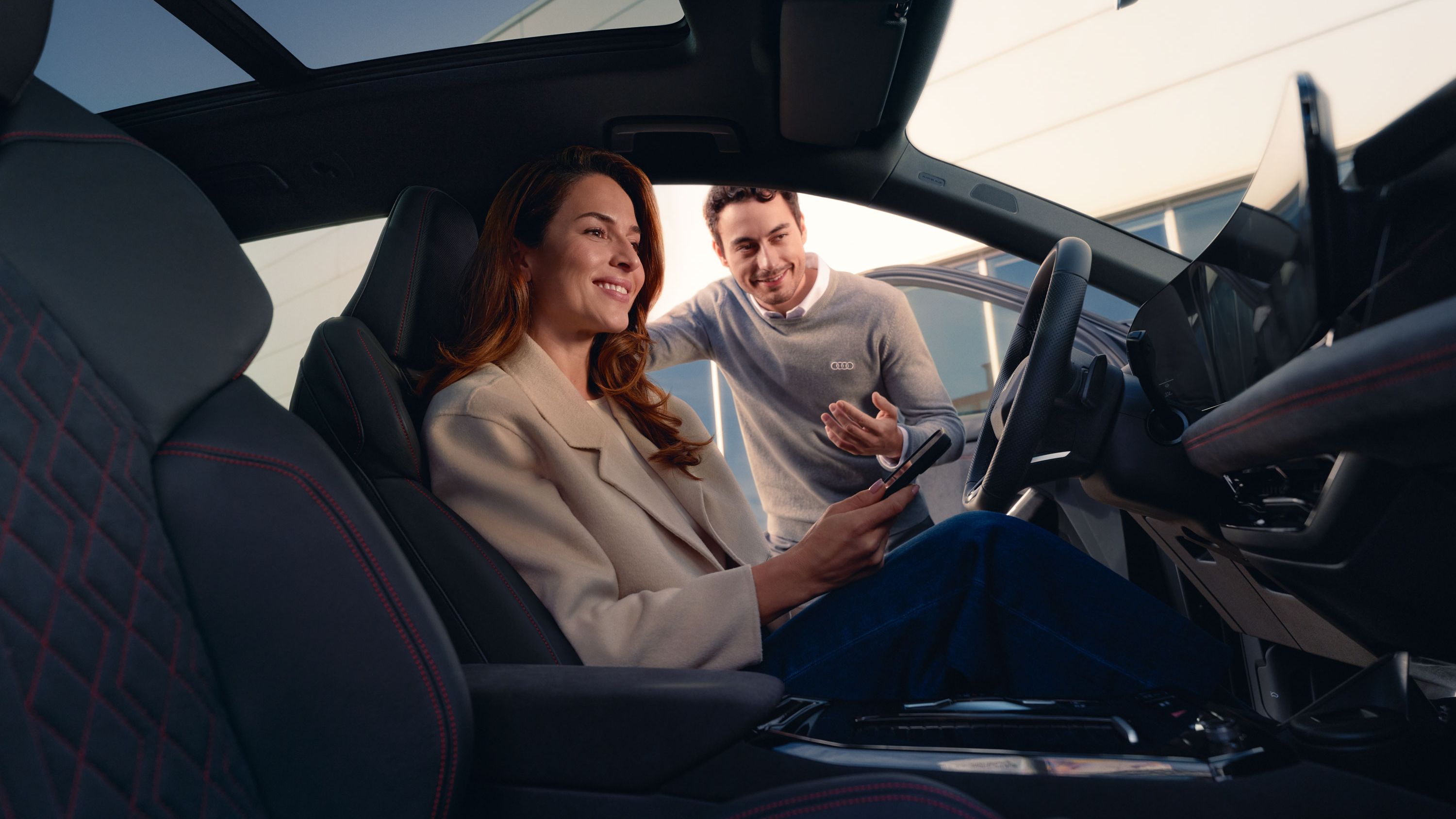 Eine Frau sitzt lächelnd auf dem Fahrersitz eines Audi und hält ein Smartphone in der Hand. Neben dem offenen Fahrzeug beugt sich ein Serviceberater herein und spricht freundlich mit ihr.
