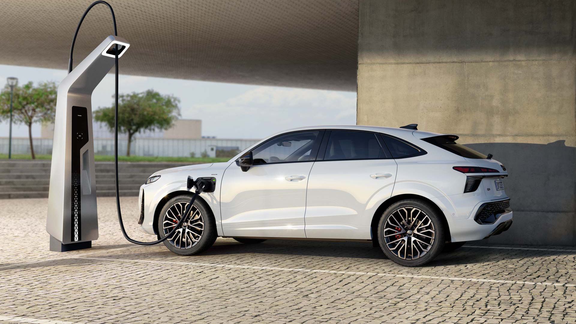 Ein weißer Audi Q3 Sportback e-hybrid lädt an einer Ladestation. Das Fahrzeug steht auf einem gepflasterten Platz unter einer großen, geometrischen Struktur. Im Hintergrund sind Bäume und ein bewölkter Himmel zu sehen.