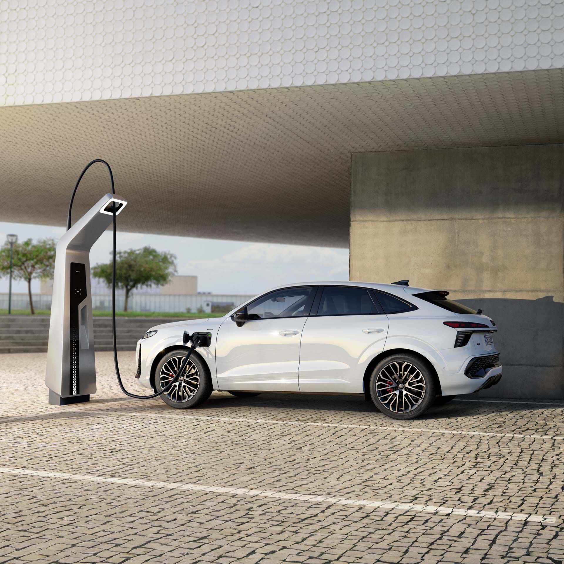 Ein weißer Audi Q3 Sportback e-hybrid lädt an einer Ladestation. Das Fahrzeug steht auf einem gepflasterten Platz unter einer großen, geometrischen Struktur. Im Hintergrund sind Bäume und ein bewölkter Himmel zu sehen.