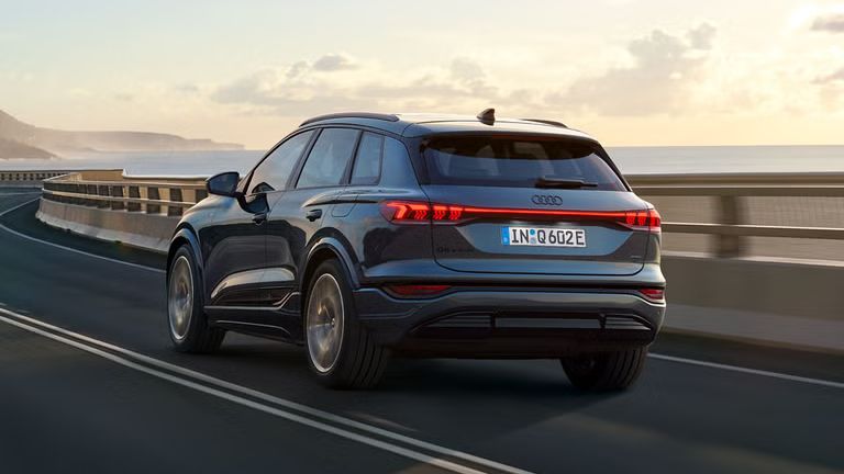 Schräge Heckaufnahme des Q6 SUV e-tron, welche über eine kurvige Straße am Meer fährt. Im Hintergrund sind Berge zu sehen.