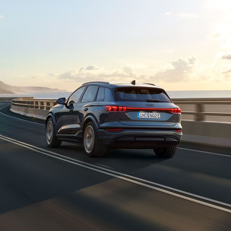 Schräge Heckaufnahme des Q6 SUV e-tron, welche über eine kurvige Straße am Meer fährt. Im Hintergrund sind Berge zu sehen.