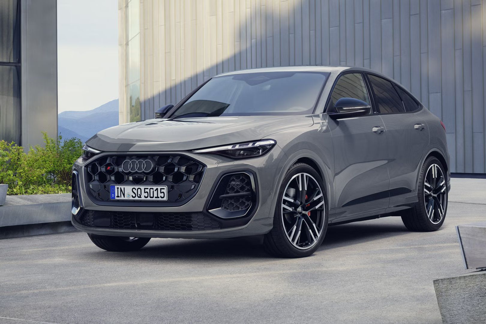 Der SQ5 Sportback steht im schrägen Frontprofil vor einem großen Gebäude. Das Fahrzeug ist Nardograu lackiert.