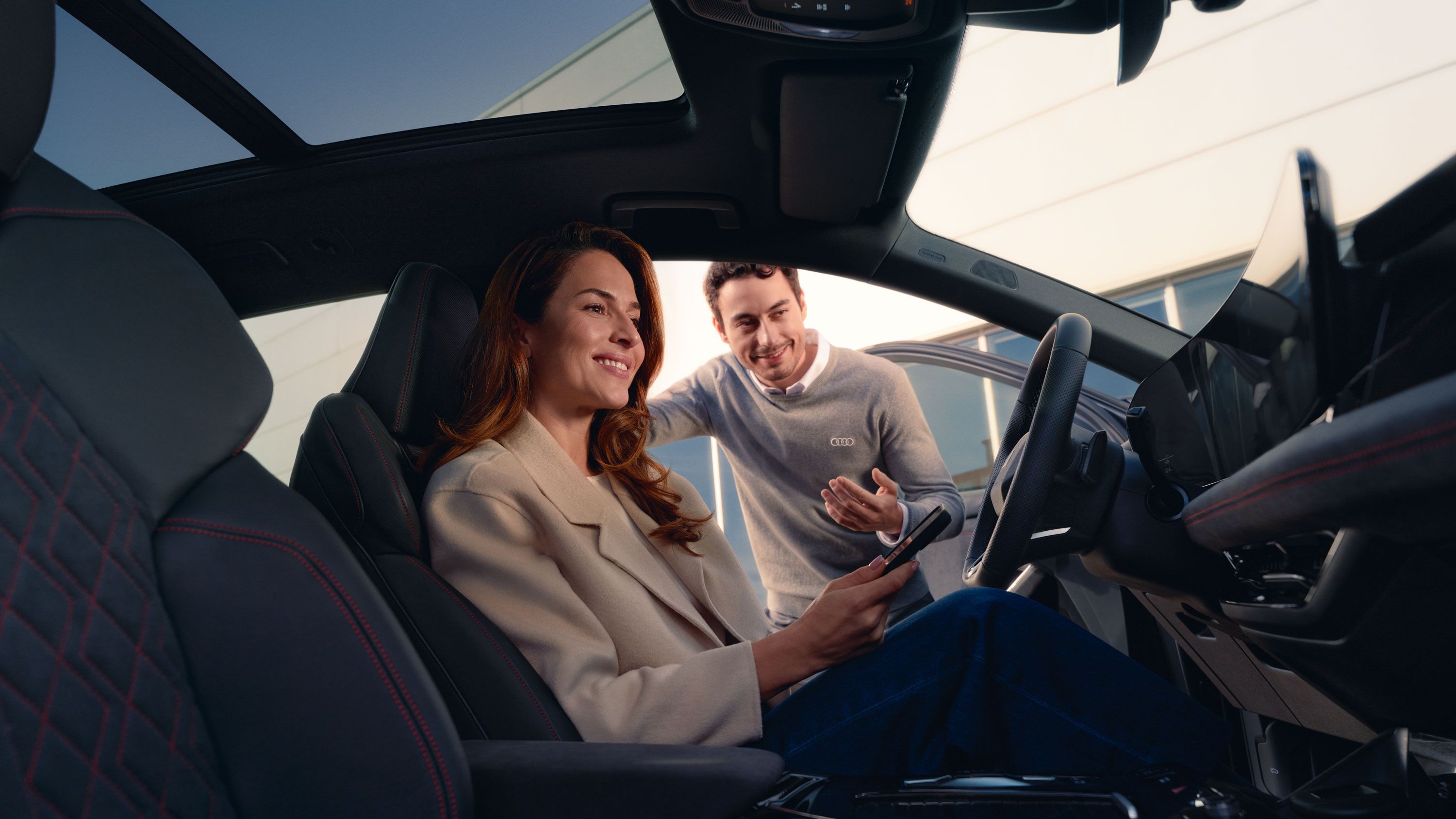 Eine Frau sitzt lächelnd auf dem Fahrersitz eines Audi und hält ein Smartphone in der Hand. Neben dem offenen Fahrzeug beugt sich ein Serviceberater herein und spricht freundlich mit ihr.