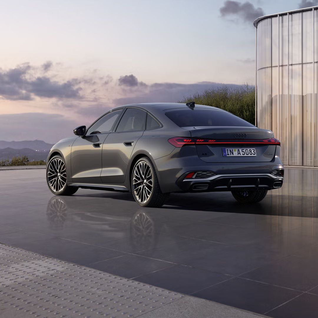 Die schwarze Edition One mit Akzenten in Magnesiumgrau der A5 Limousine in schrägen Heckprofil spiegelt sich in dem glatten Untergrund. Im Hintergrund sind Berge bei Dämmerung zu sehen.