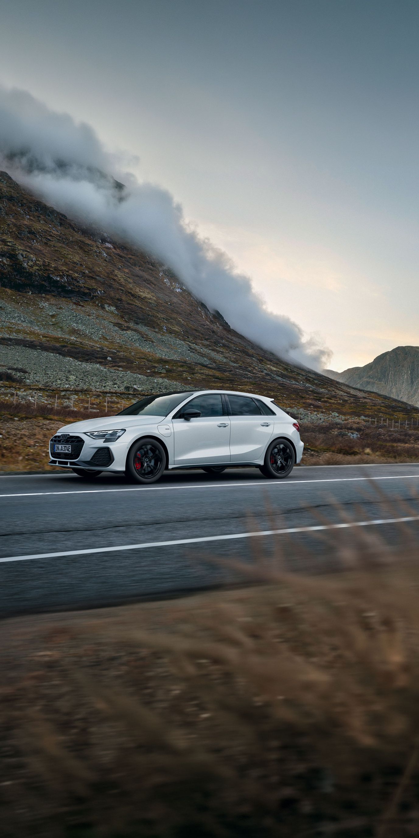 Weißer Audi A3 Sportback e-hybrid fährt auf einer leeren Landstraße durch eine bergige Landschaft. Links erhebt sich ein steiler, felsiger Hang, aus dem sich Nebelschwaden lösen. Die Stimmung ist herbstlich und ruhig, mit gedämpften Farben und bewölktem Himmel.