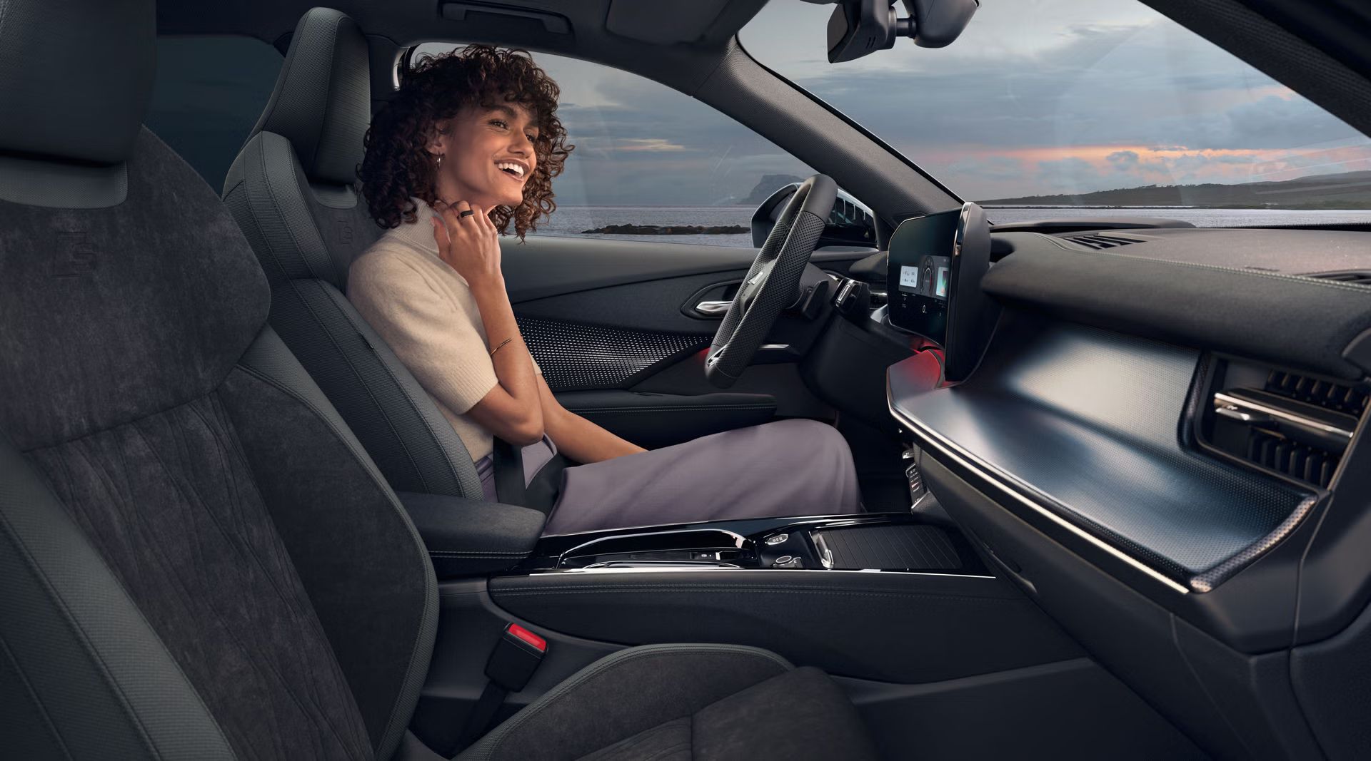Vorderer Innenraum des Q3 SUV mit erhöhten Sitzen und Panoramadisplay. Auf der Fahrerseite sitzt eine junge, modisch gekleidete Frau und lächelt.