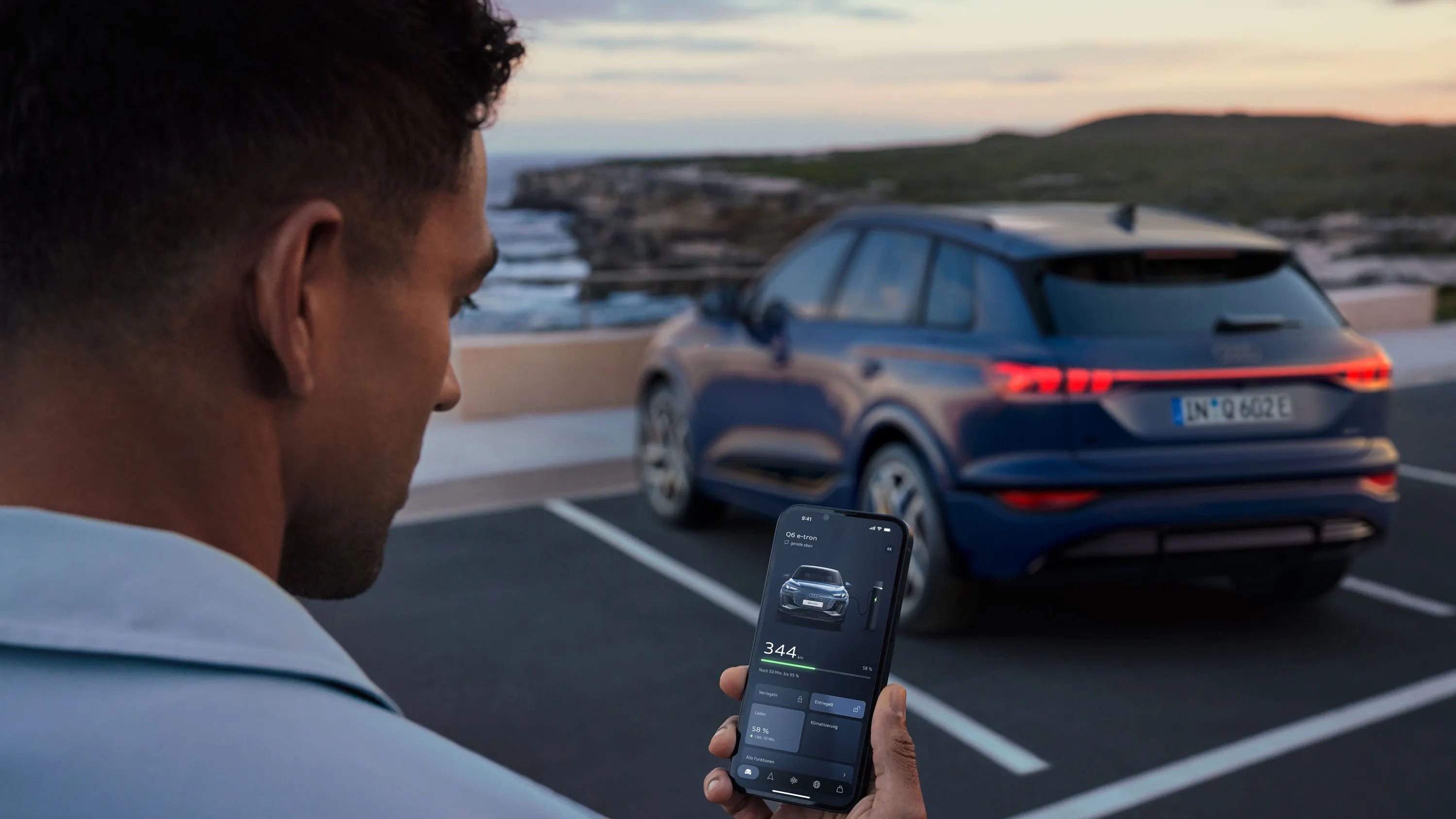 Ein Mann steht auf einem Parkplatz und hält ein Smartphone in der Hand. Im Hintergrund steht ein blauer Audi auf einem Stellplatz vor einer Küstenlandschaft. 