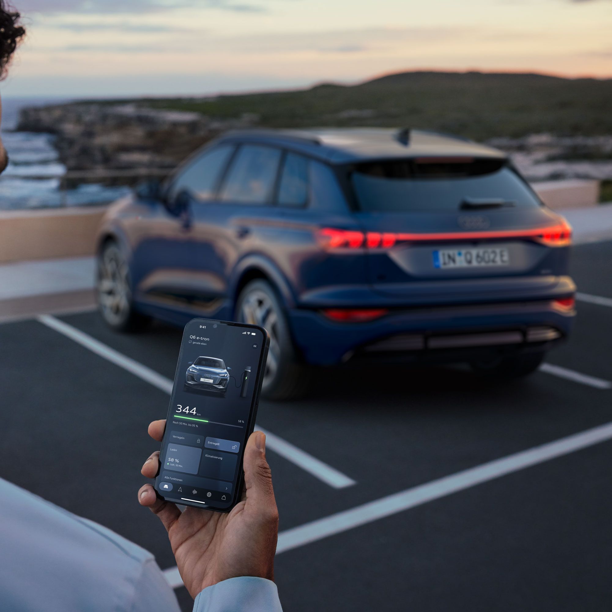 Ein Mann steht auf einem Parkplatz und hält ein Smartphone in der Hand. Im Hintergrund steht ein blauer Audi auf einem Stellplatz vor einer Küstenlandschaft. 