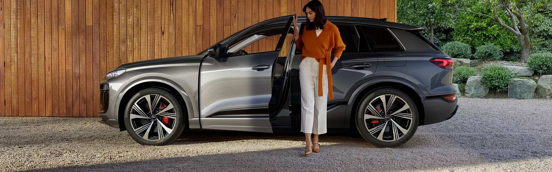 Der SQ6 SUV e-tron steht seitlich vor einer Holzhausfassade, während sich eine junge Frau an der offenen Fahrertür festhält. Auf der rechten Seite des Bildes ist der Vorgarten des Hauses zu sehen.