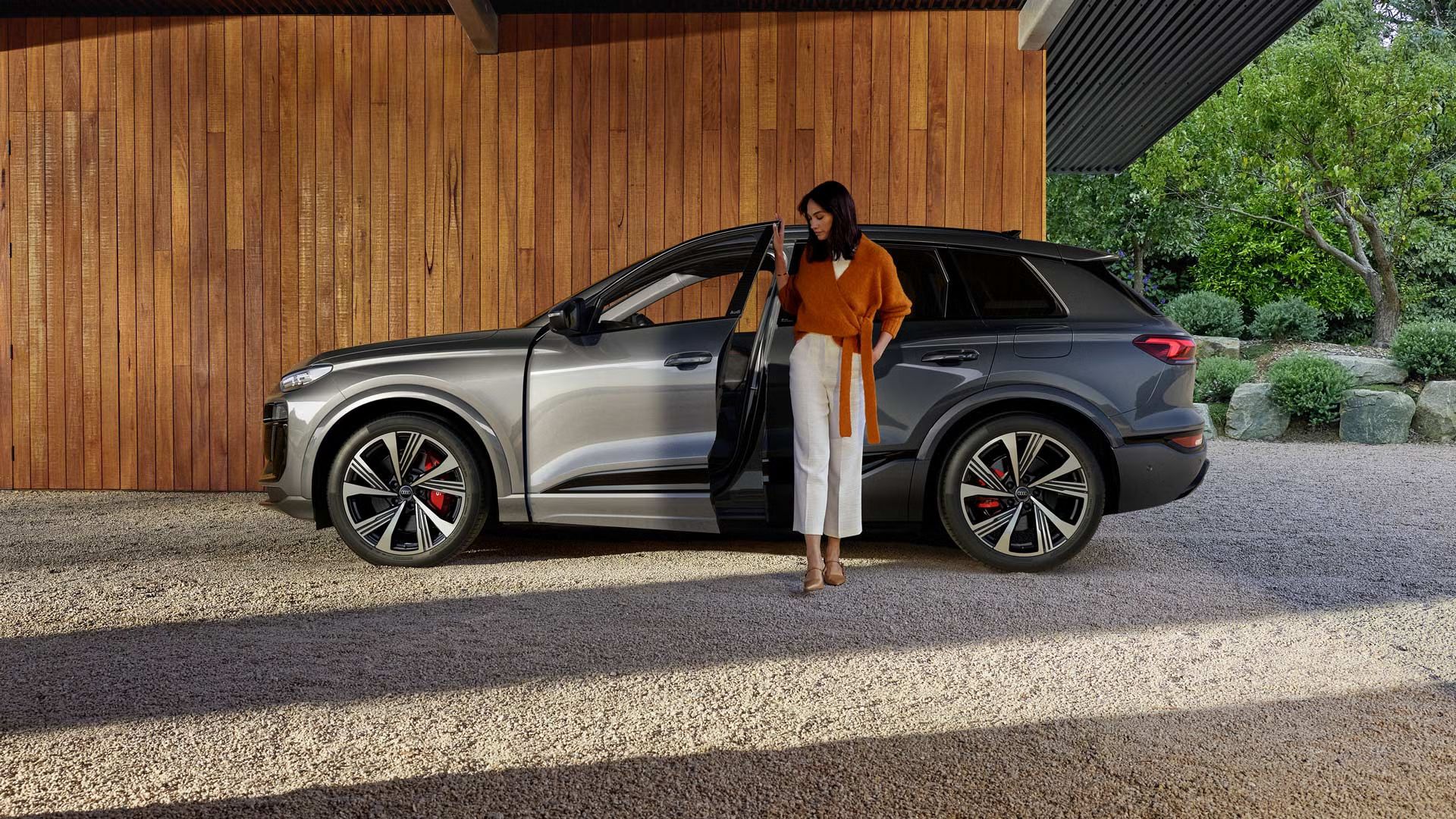 Der SQ6 SUV e-tron steht seitlich vor einer Holzhausfassade, während sich eine junge Frau an der offenen Fahrertür festhält. Auf der rechten Seite des Bildes ist der Vorgarten des Hauses zu sehen.