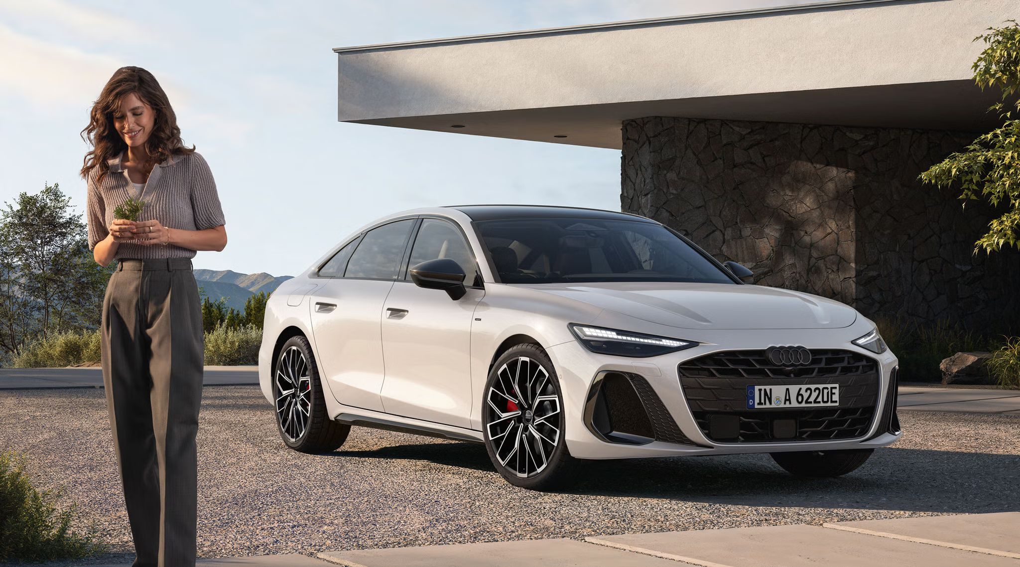 Die A6 Limousine e-hybrid steht vor einem Haus. Vor dem Auto steht eine junge Frau.